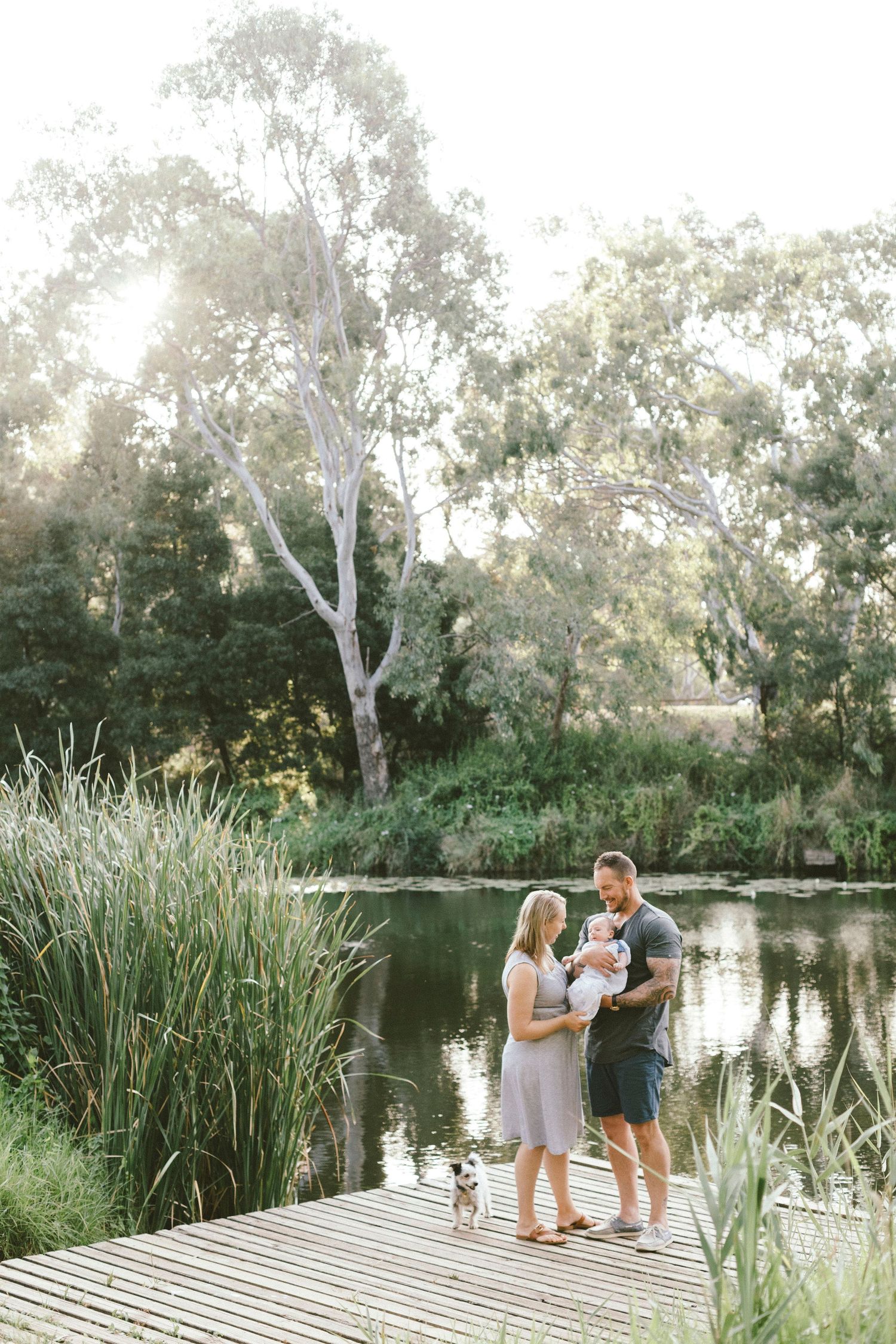 A family shares an intimate moment on a wooden dock by a peaceful pond surrounded by trees.