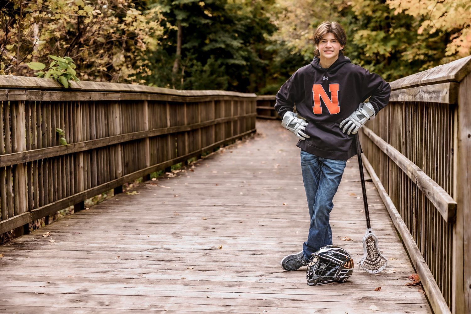 Student athlete in dark sweatshirt posing with lacrosse stick on wooden bridge during autumn.