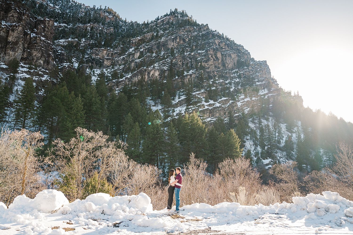 A couple stands embracing against a snowy mountain backdrop with dramatic winter lighting.