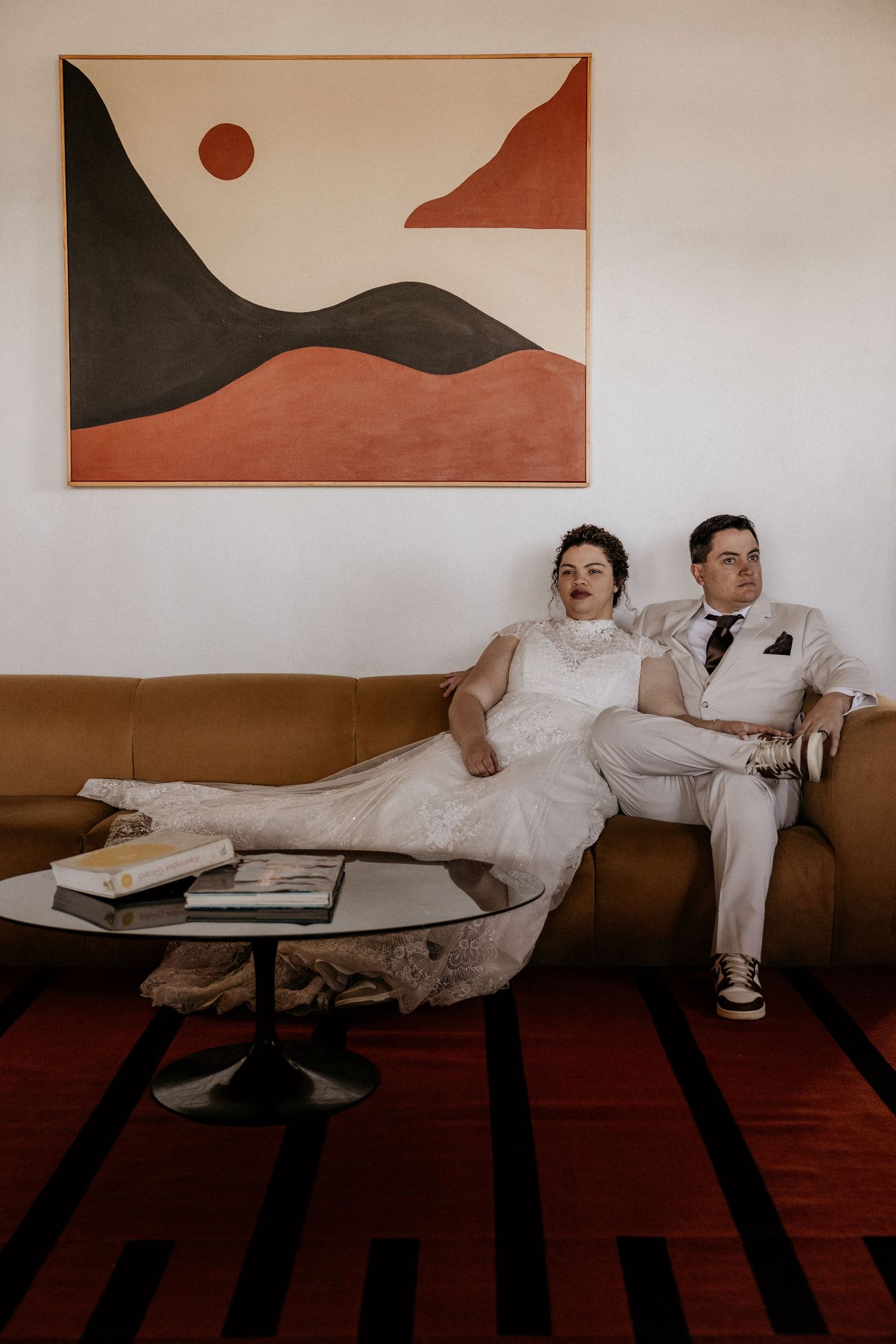 Bride and groom relaxing on tan leather sofa beneath abstract mid-century modern wall art painting.