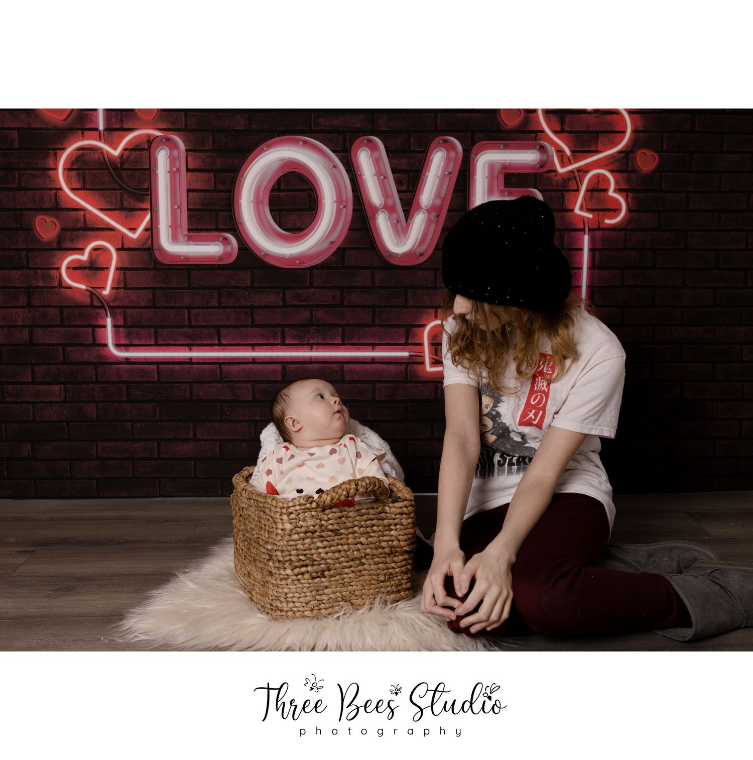 Studio portrait with neon love sign and baby in woven basket against brick wall backdrop.