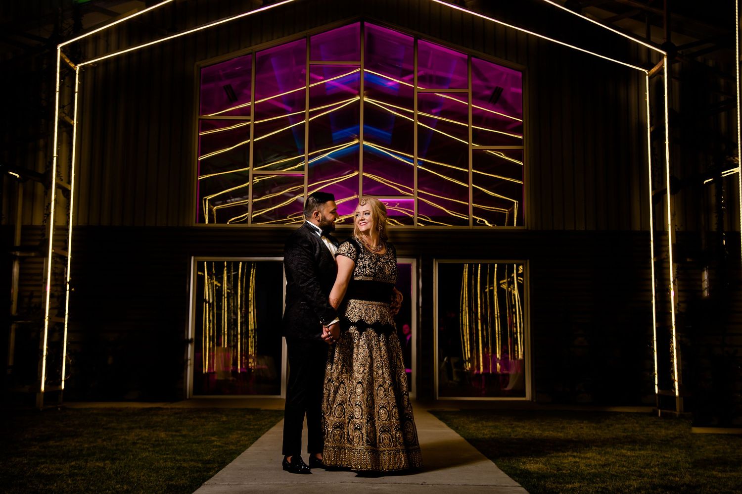 Couple poses at night in front of illuminated barn-style venue with geometric lighting.