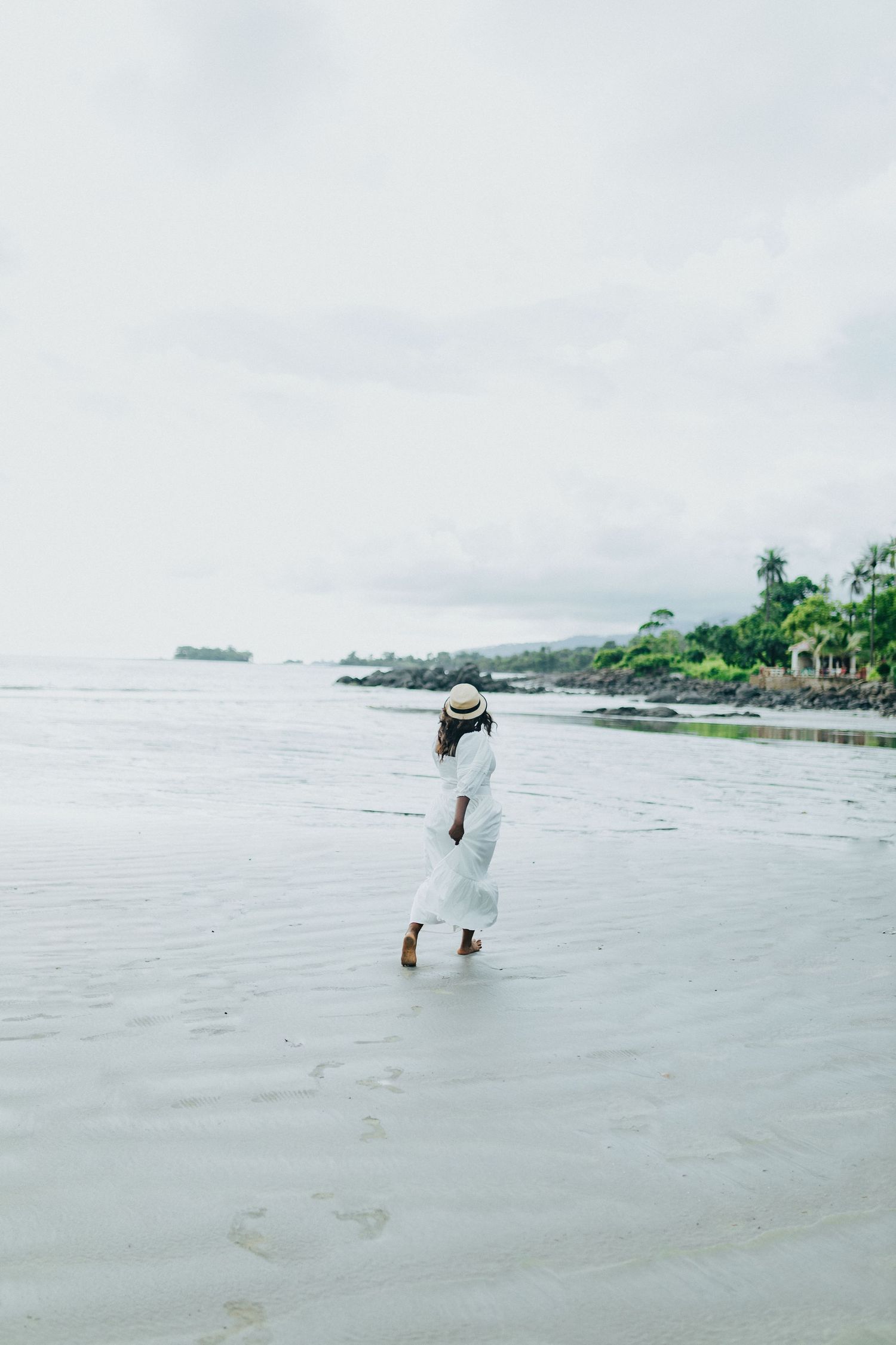 A solitary figure in white dress walks along a misty beach shore with palm trees in the distance.
