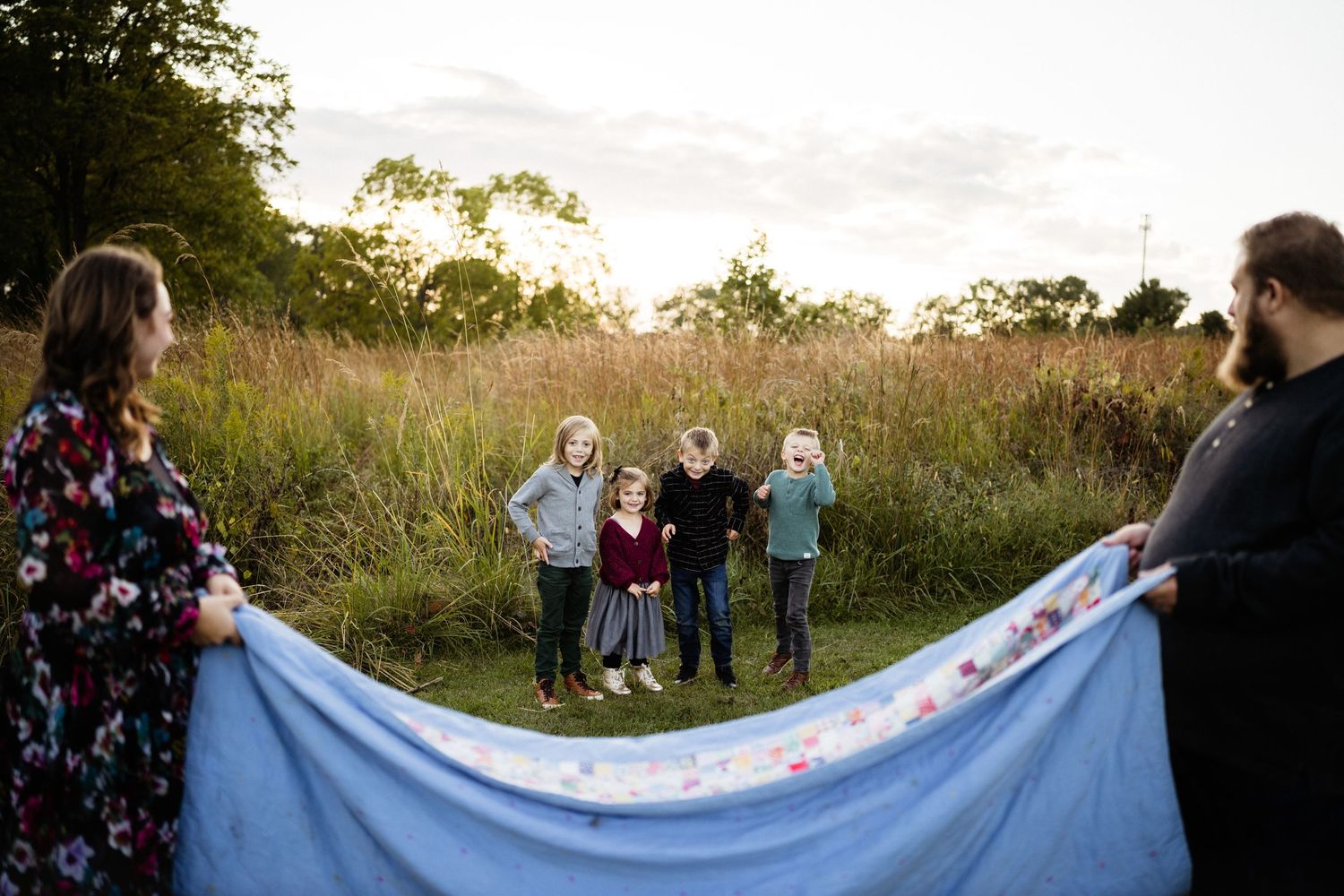 Two people hold blanket while others stand in field with sunset in background.