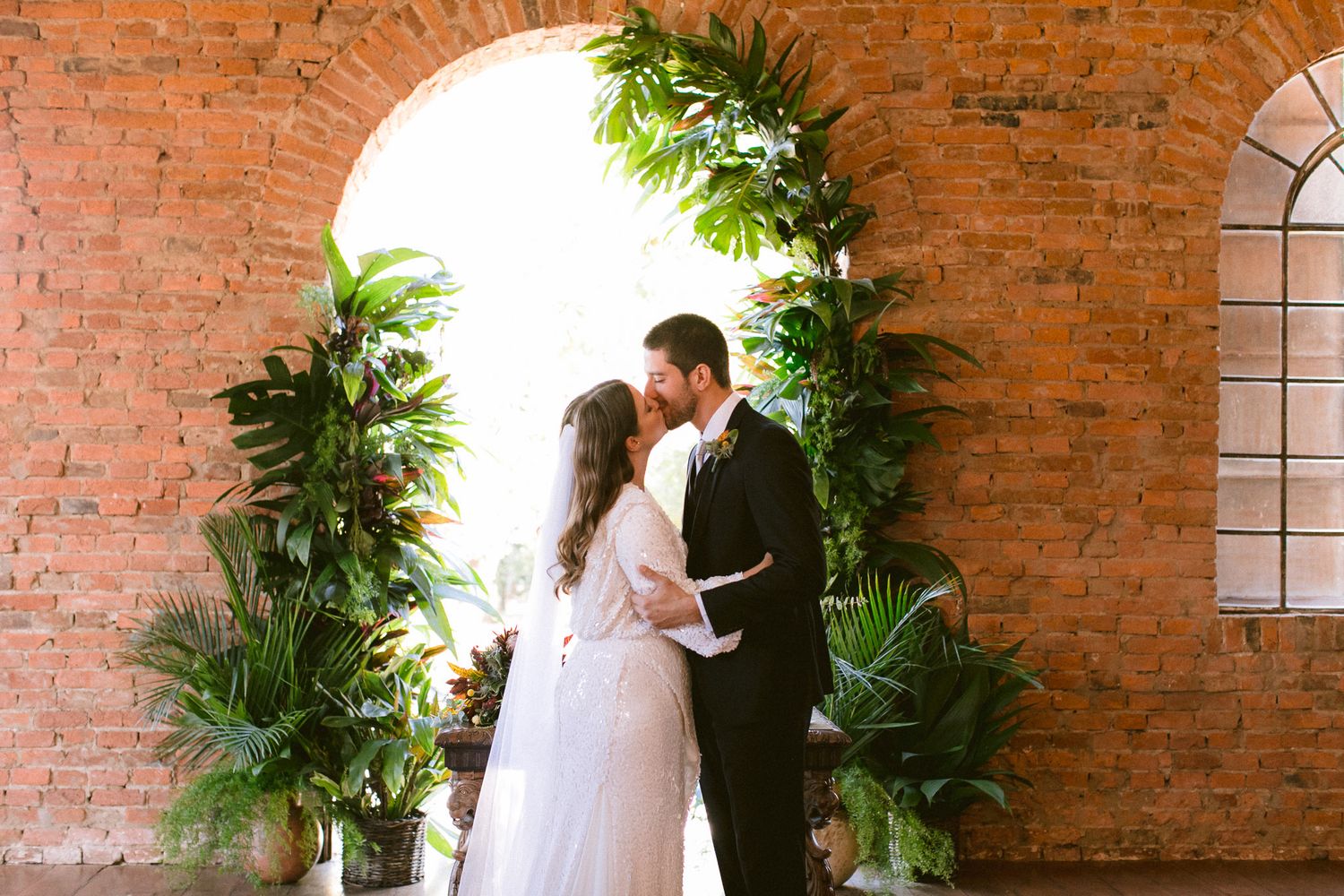Wedding couple embracing under brick archway decorated with tropical greenery.