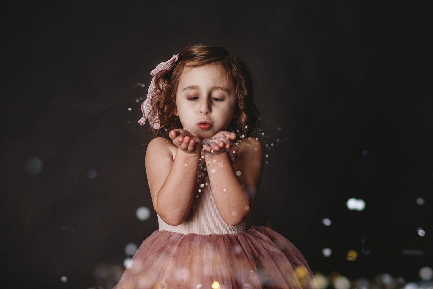 Young child in a pink tulle dress blowing glitter in a dark studio setting.