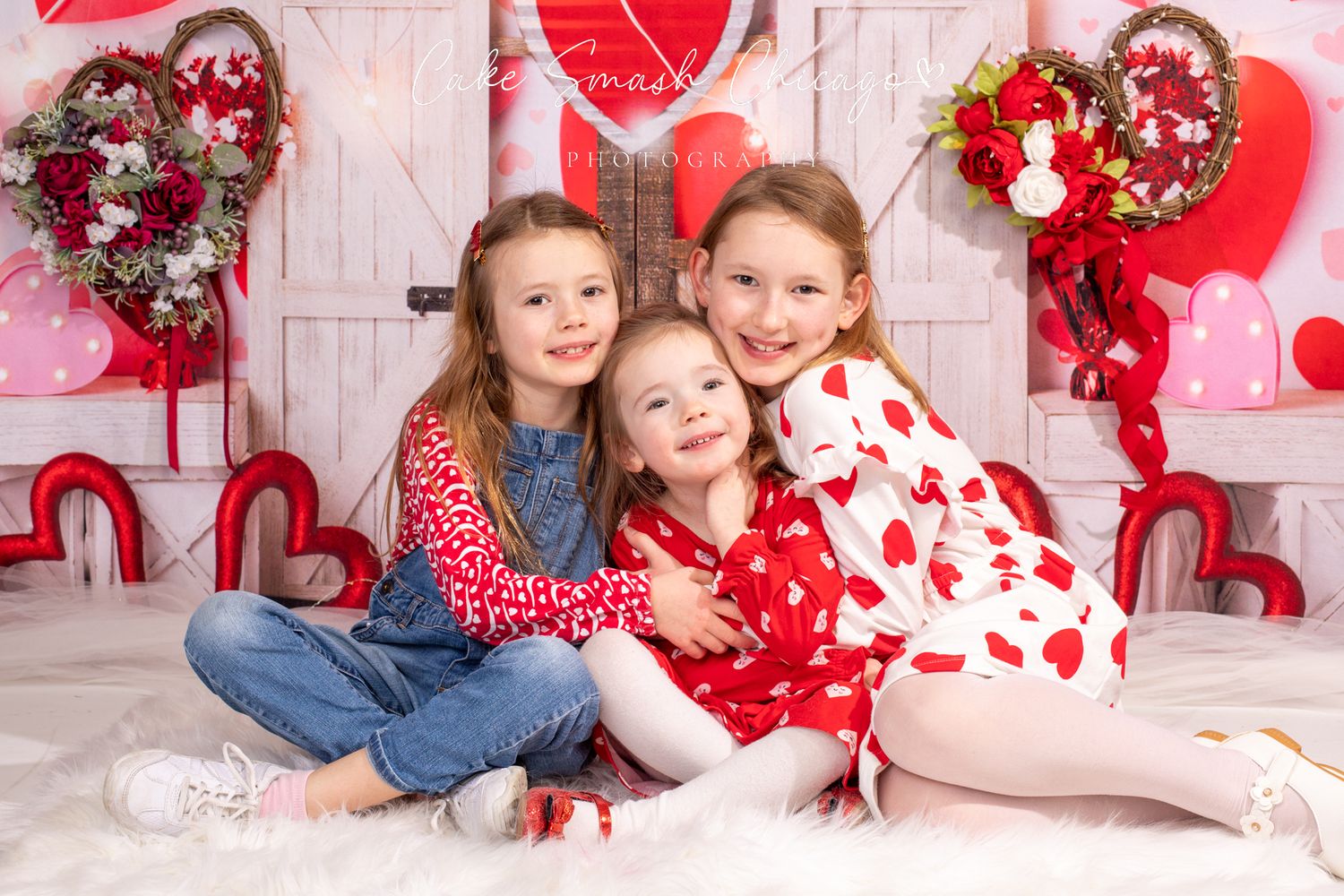 Three children in Valentine's themed outfits sitting together against a heart-decorated background.