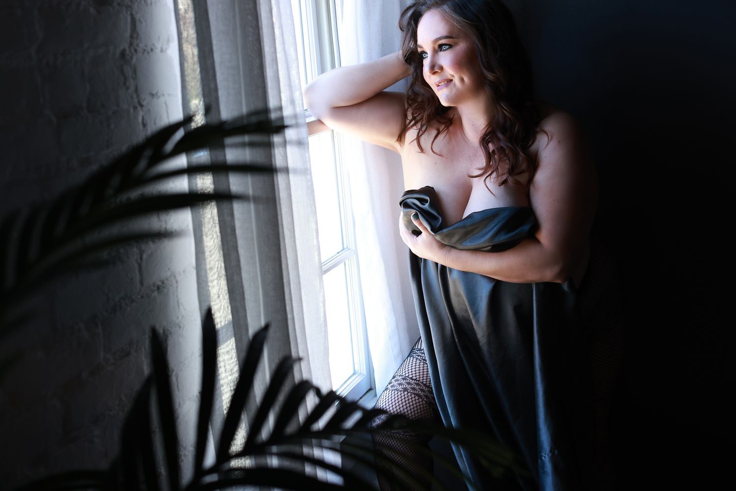 A figure in a dark dress stands by a bright window holding a cup while palm frond shadows create dramatic lighting.