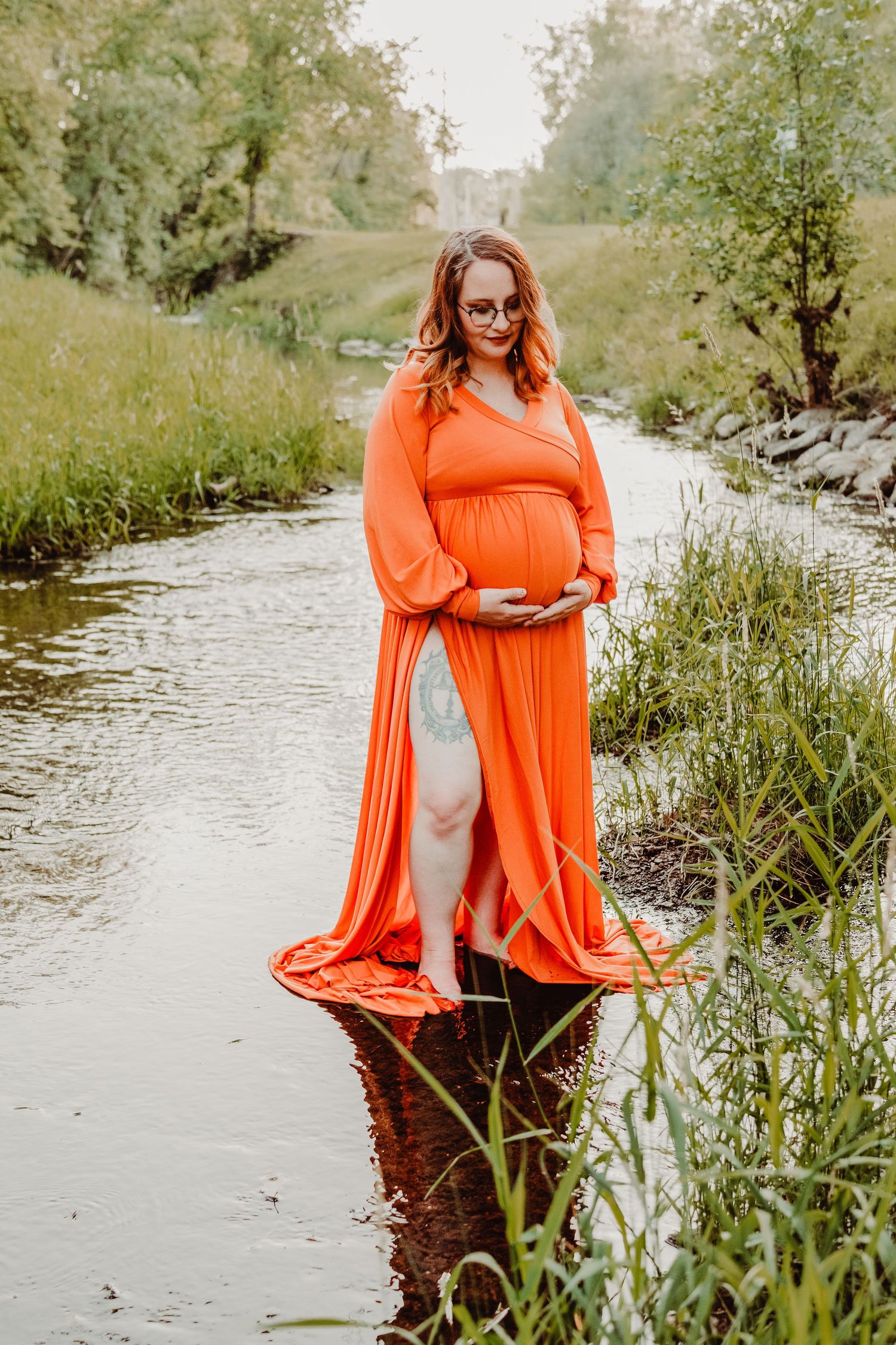 A flowing orange maternity dress reflects beautifully in a calm stream surrounded by lush greenery.