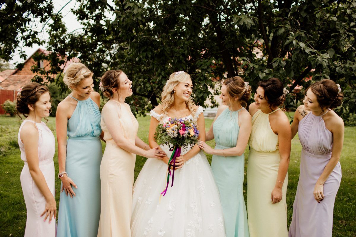 Blonde bride in lace gown holding bouquet surrounded by six bridesmaids in pastel dresses.