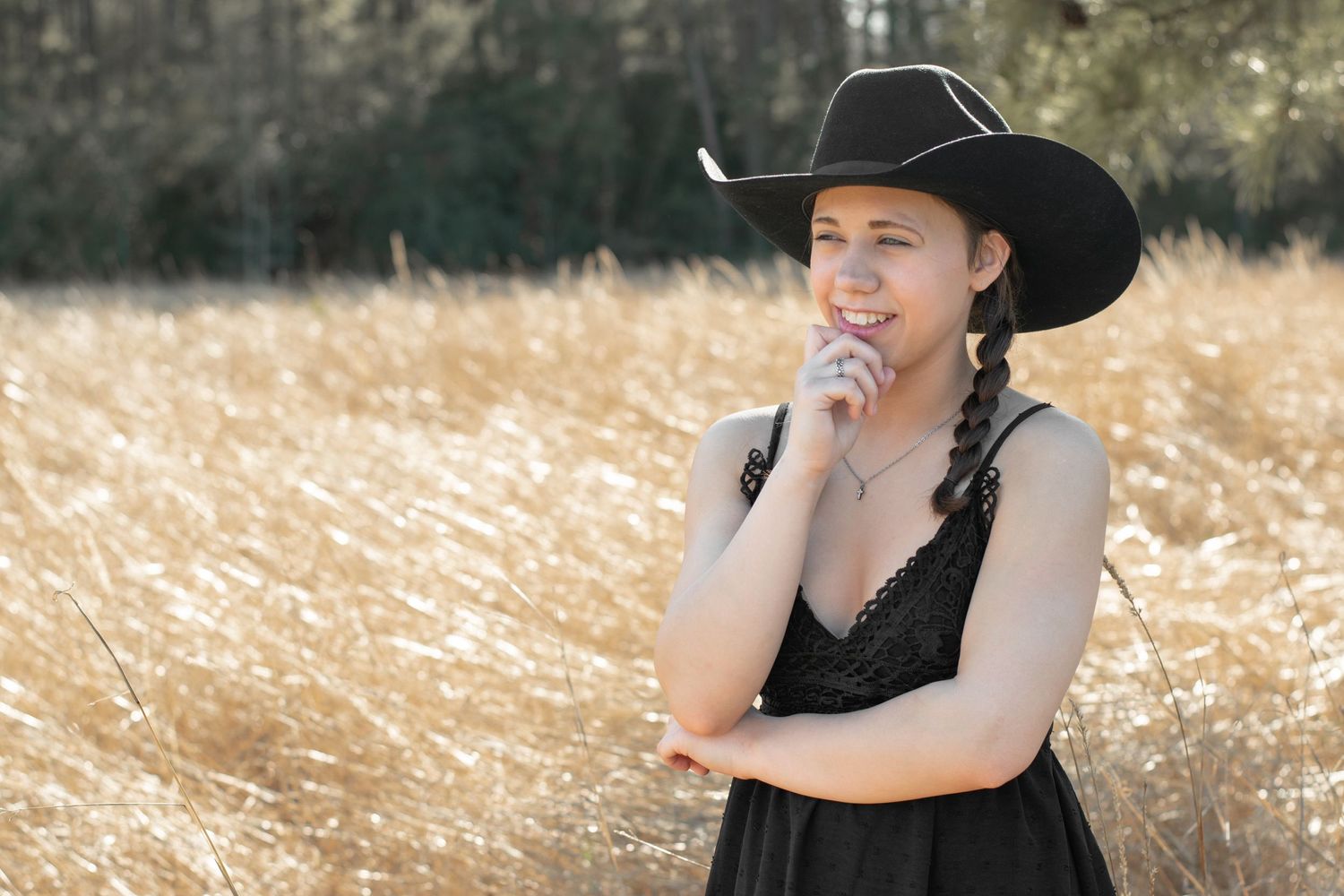 Person in black dress and cowboy hat poses thoughtfully in sunlit field.