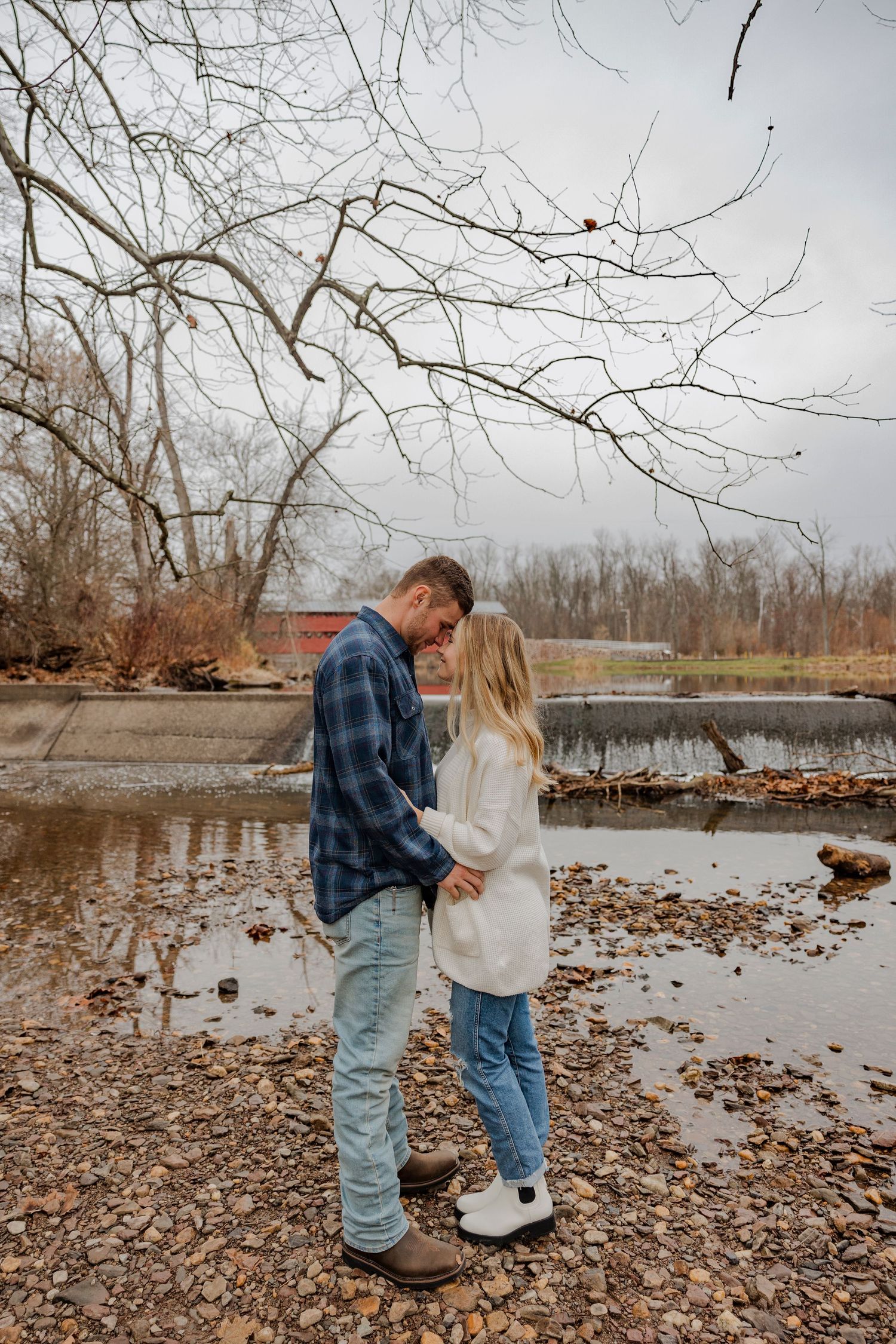 Intimate couple's portrait by a riverside with bare winter trees in the background.