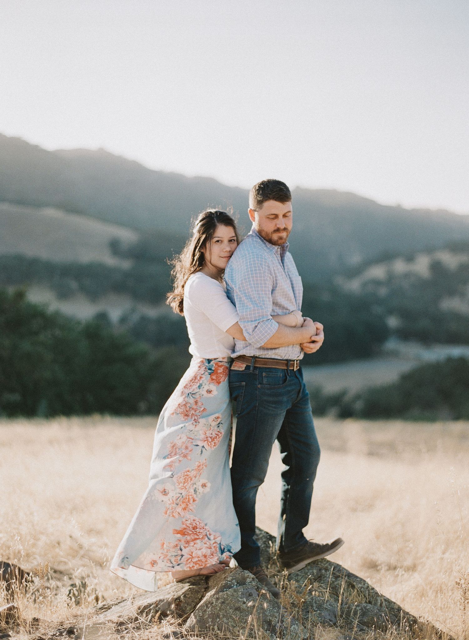 Couple embracing on rocky outcrop overlooking scenic mountain valley at sunset.