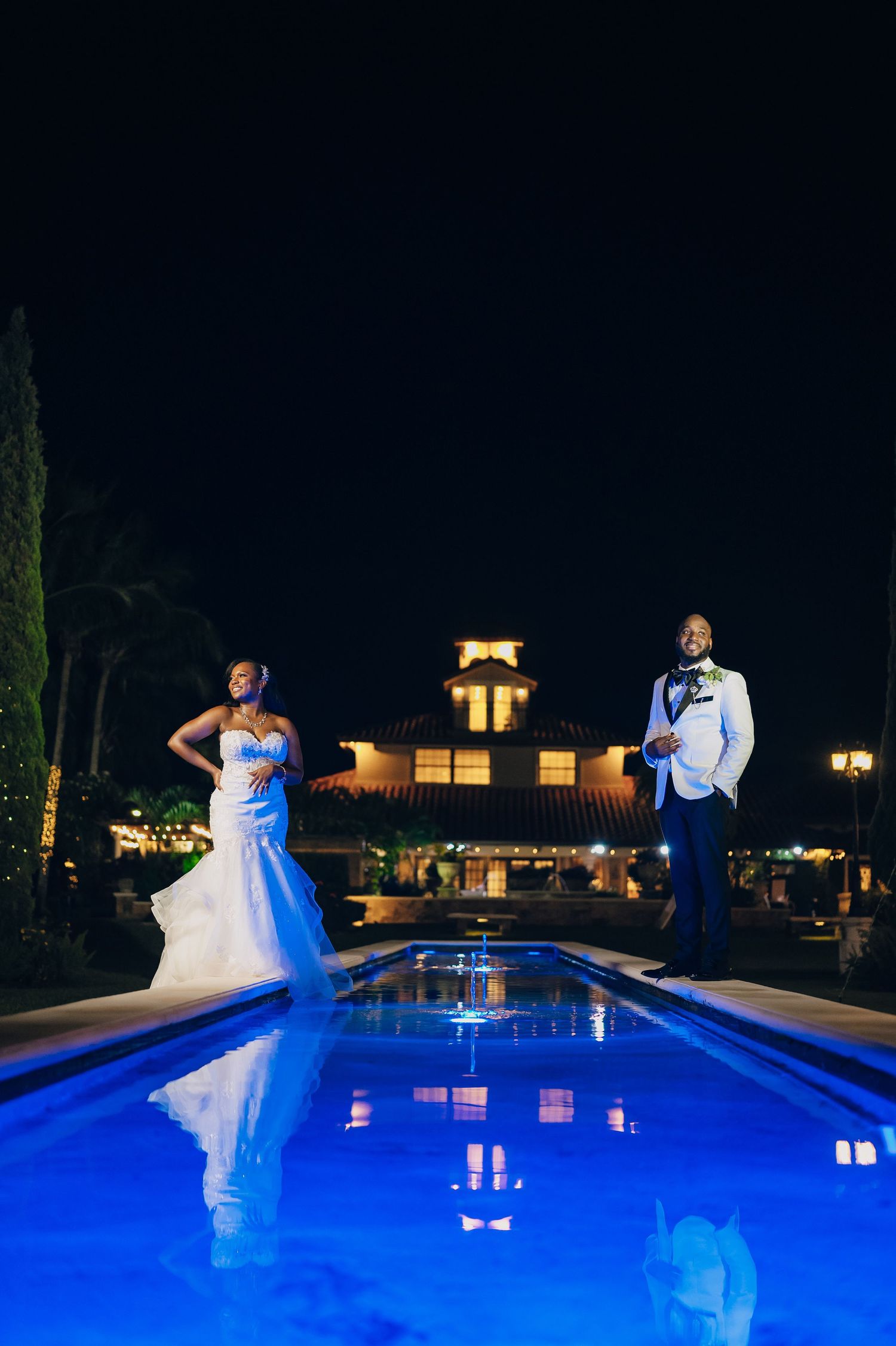 Elegant couple poses by illuminated blue swimming pool at nighttime luxury venue with glowing building in background.