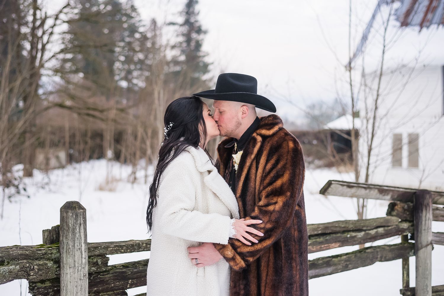 A couple kisses outside wearing winter wedding attire on their snowy, magical January wedding day. Snow covered barn in the background.