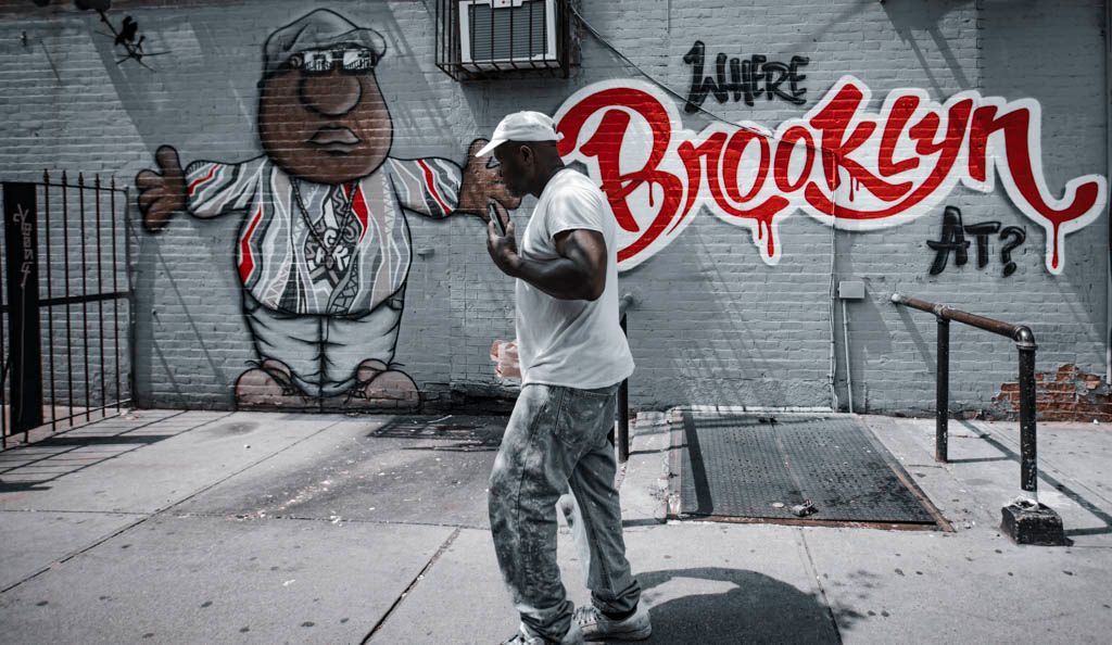 Street artist painting a colorful Brooklyn graffiti mural on a gray wall in an urban setting.