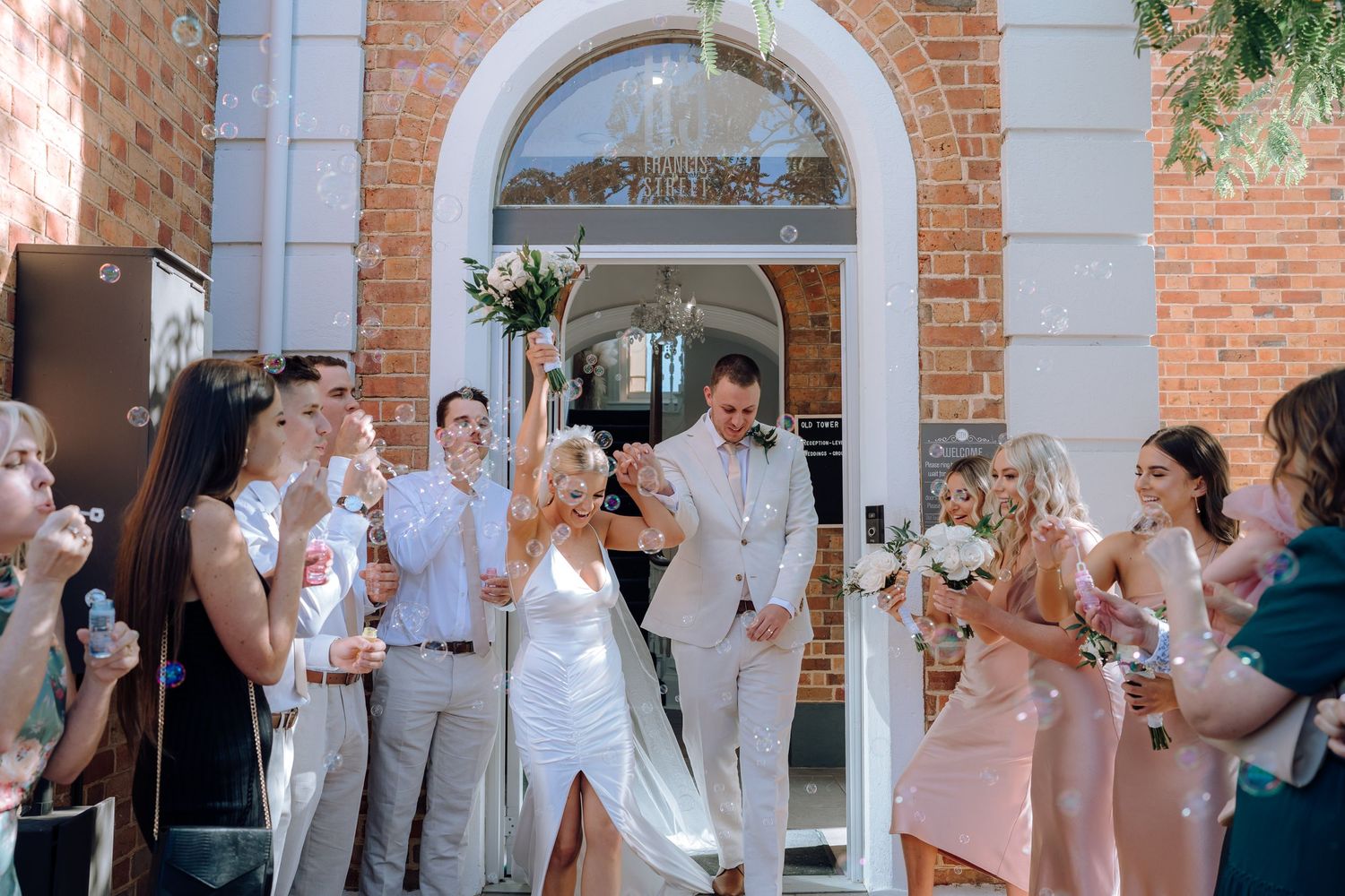 Wedding guests blow bubbles as newlyweds exit through a brick archway entrance.