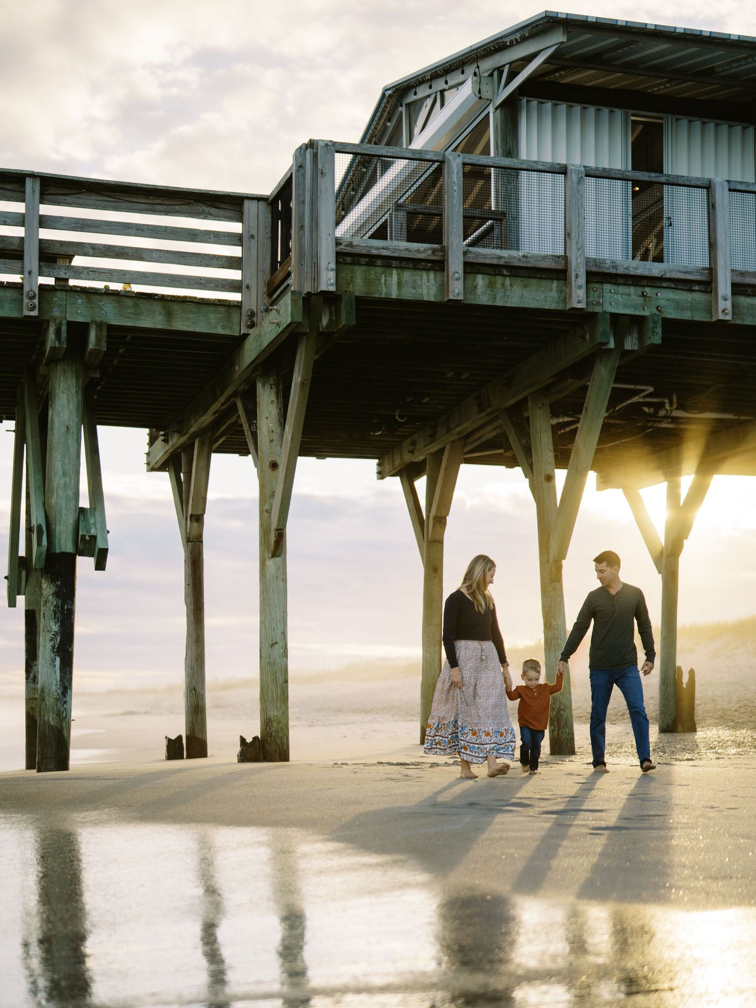 A family walks together under a wooden pier at sunset casting long shadows on the beach.