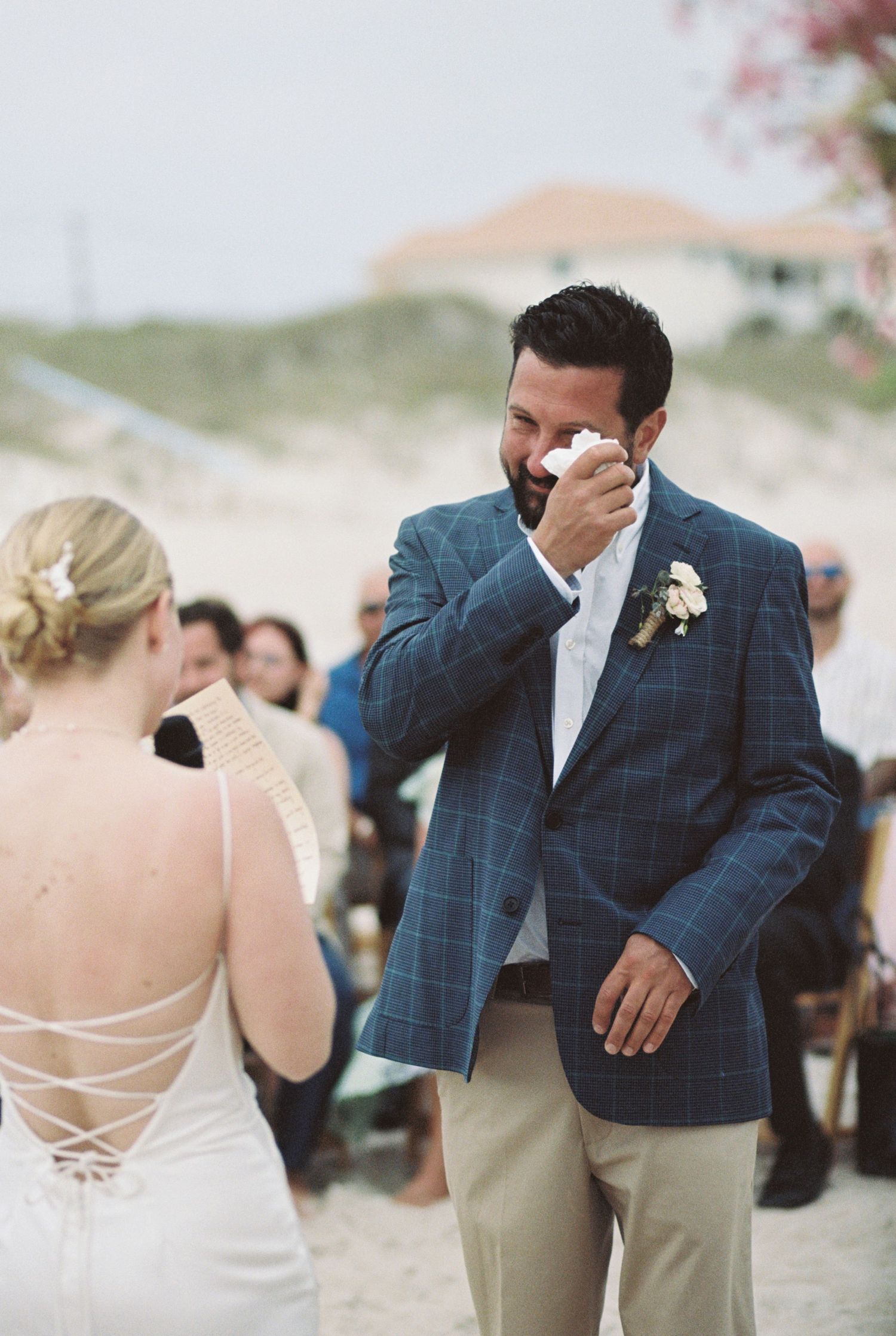 Emotional groom wipes tears during beach wedding ceremony while wearing navy checkered blazer.