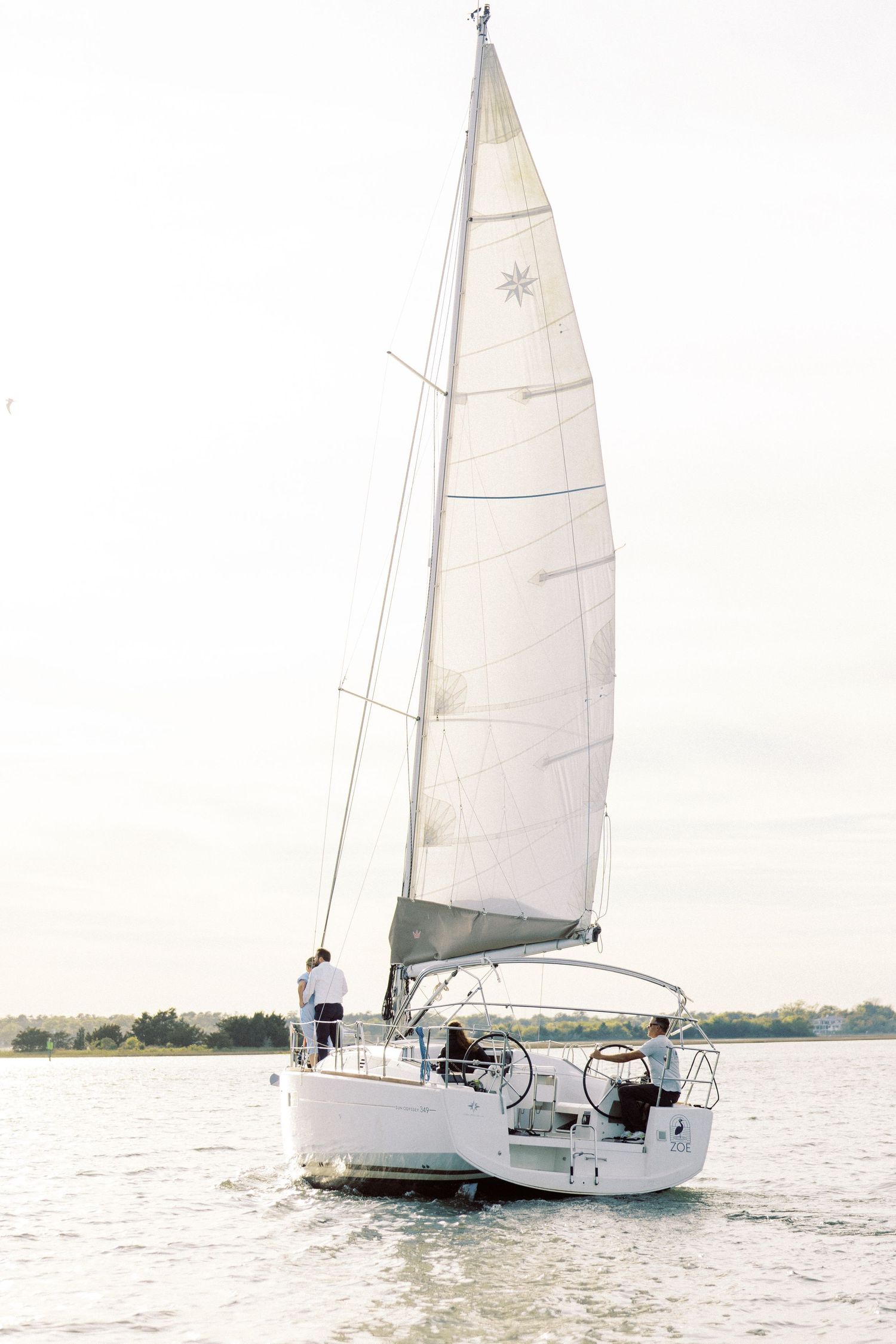 A white sailboat with raised sails glides across calm waters near the shoreline on a bright day.