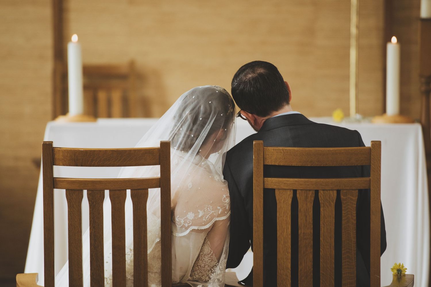 catholic bride in lace veil leans over to speak with groom in dark blue suit while seated at the alter during mass.