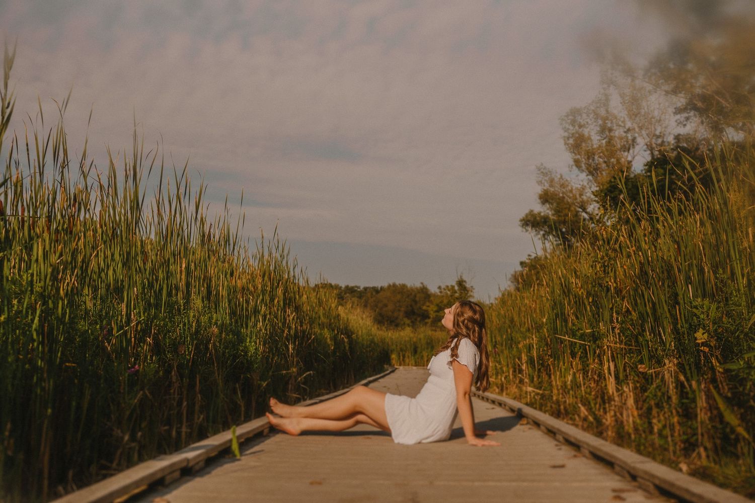 A person in a white dress reclines on a wooden boardwalk path surrounded by tall grass during golden hour.