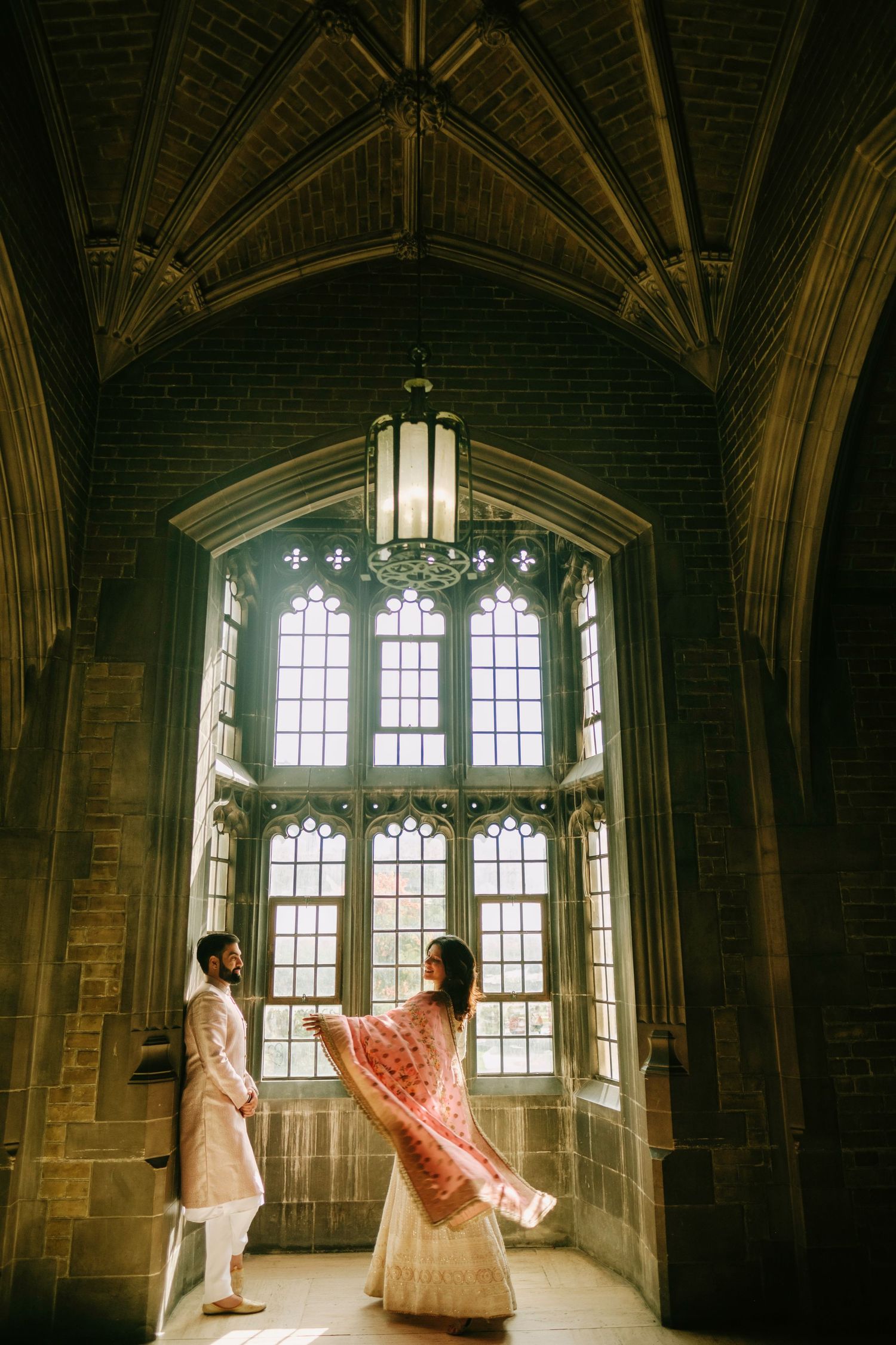 A couple stands in a grand Gothic architectural space with tall arched windows and vaulted ceilings casting dramatic light.