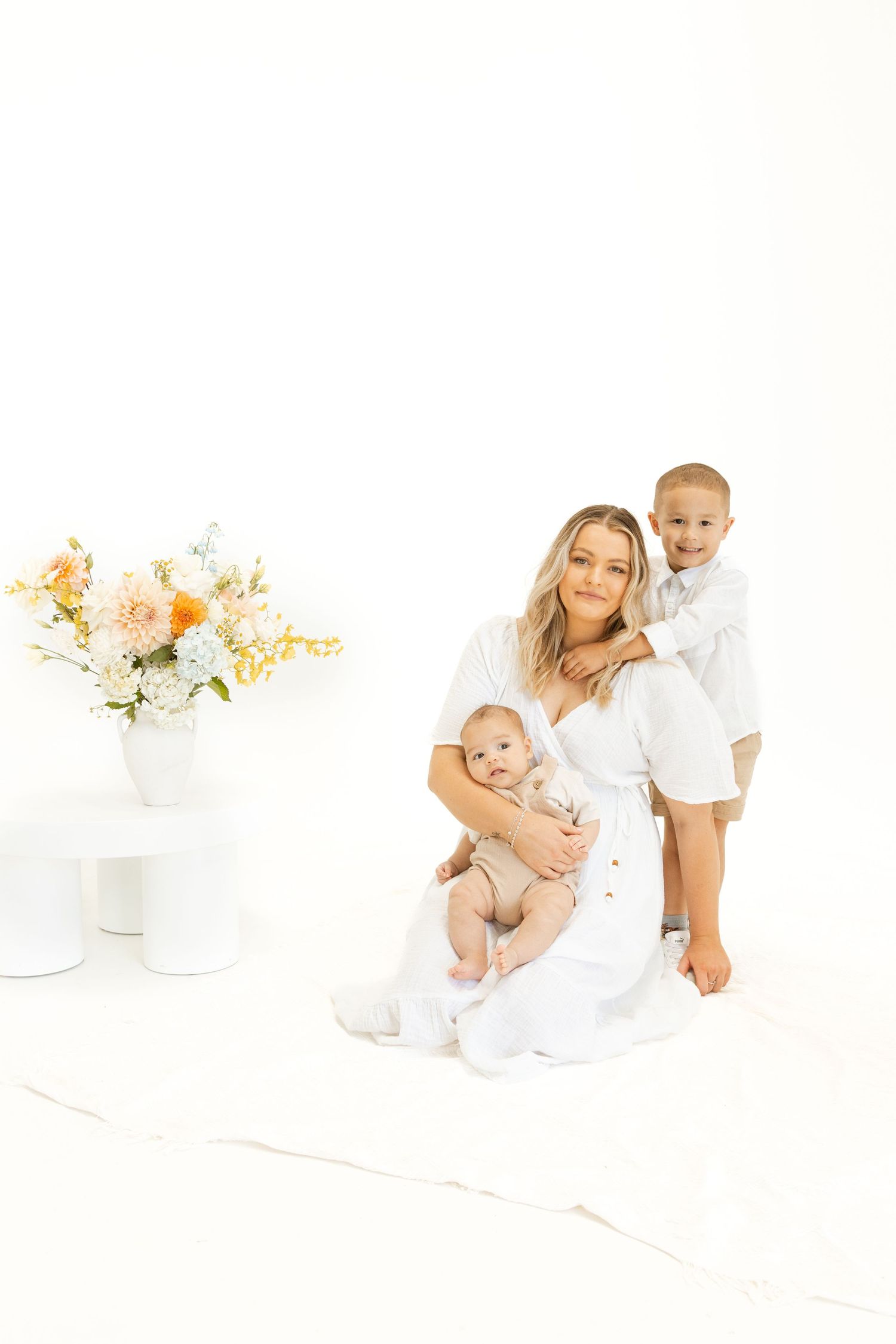 Mother in white dress poses with two children against minimalist backdrop with peachy floral arrangement.