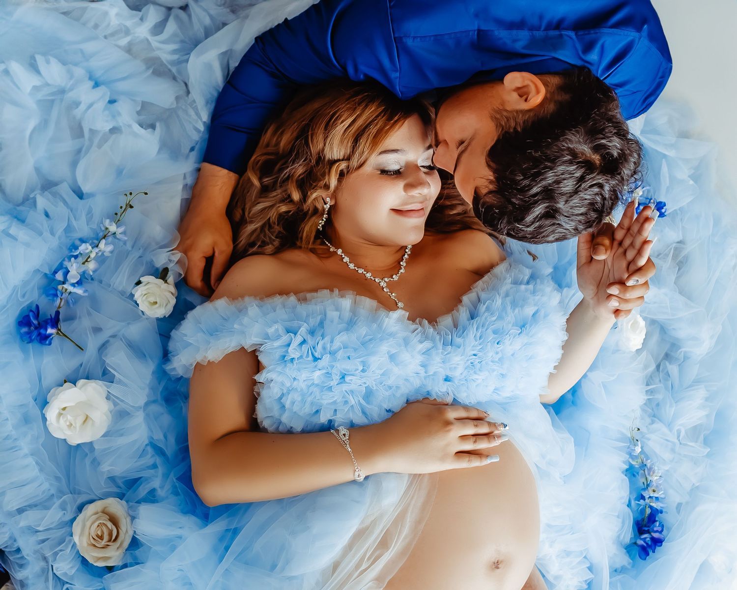 Maternity photo with couple in blue lying on flowers and fabric.