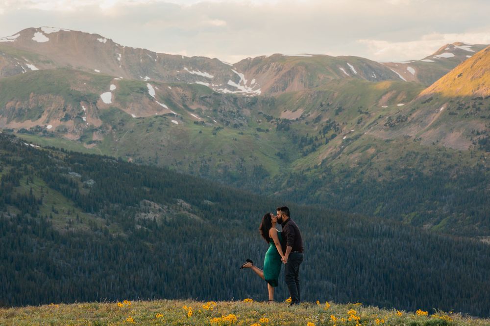 A couple embraces on a mountain summit with snow-capped peaks and valleys in the background.