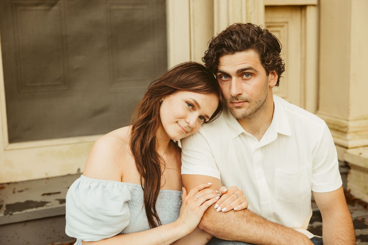 A romantic summer portrait of a couple in light clothing against a rustic building backdrop.