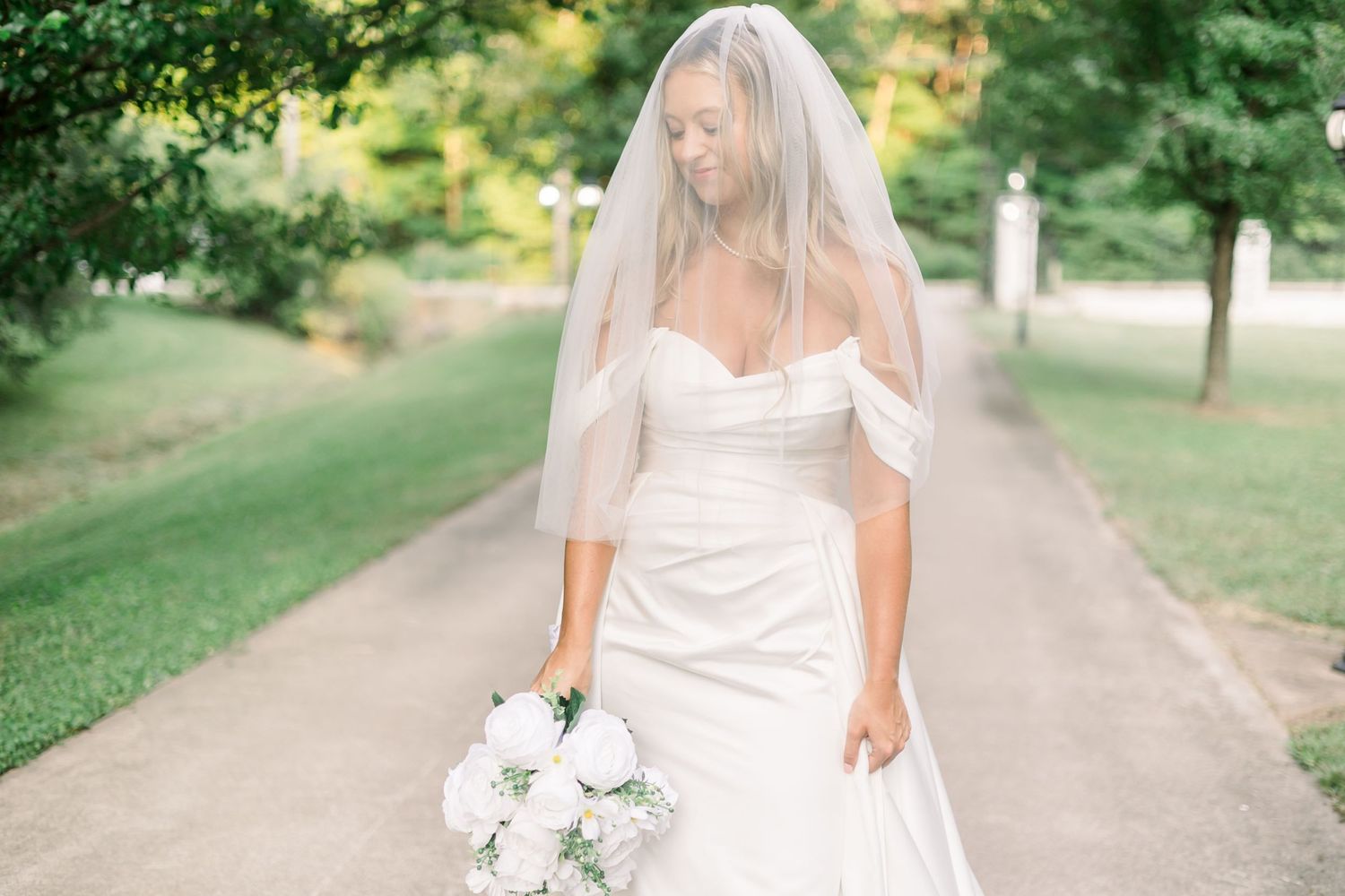 Elegant bride in flowing white dress poses in a sunlit garden pathway.