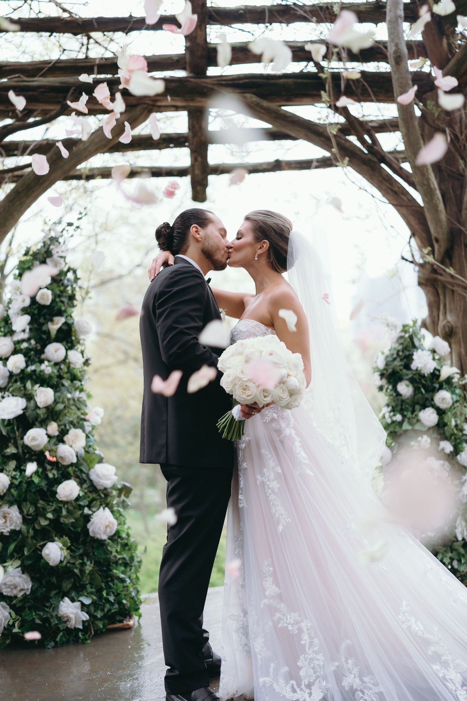 Romantic wedding kiss under flower-adorned wooden arch with falling petals.