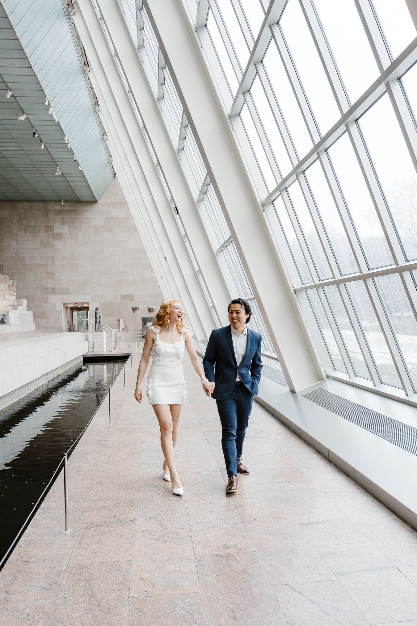 A couple walks through a modern architectural space with slanted glass windows and sleek marble floors at a wedding venue.