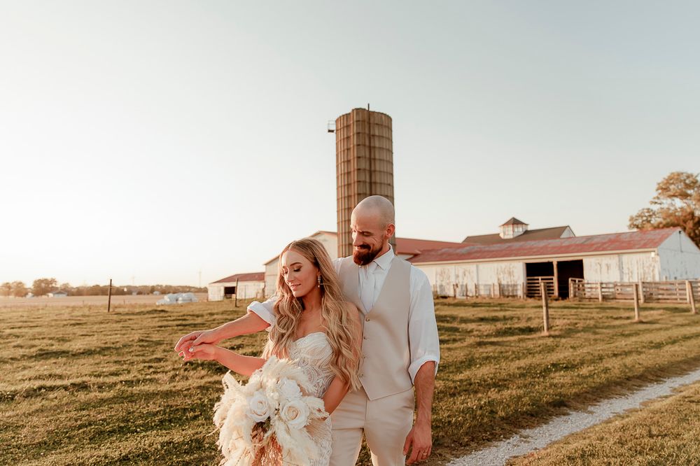 A couple in white outfits walking together on a path near a rustic barn during golden hour sunset.