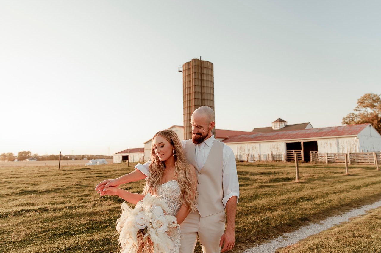 A couple in white outfits walking together on a path near a rustic barn during golden hour sunset.