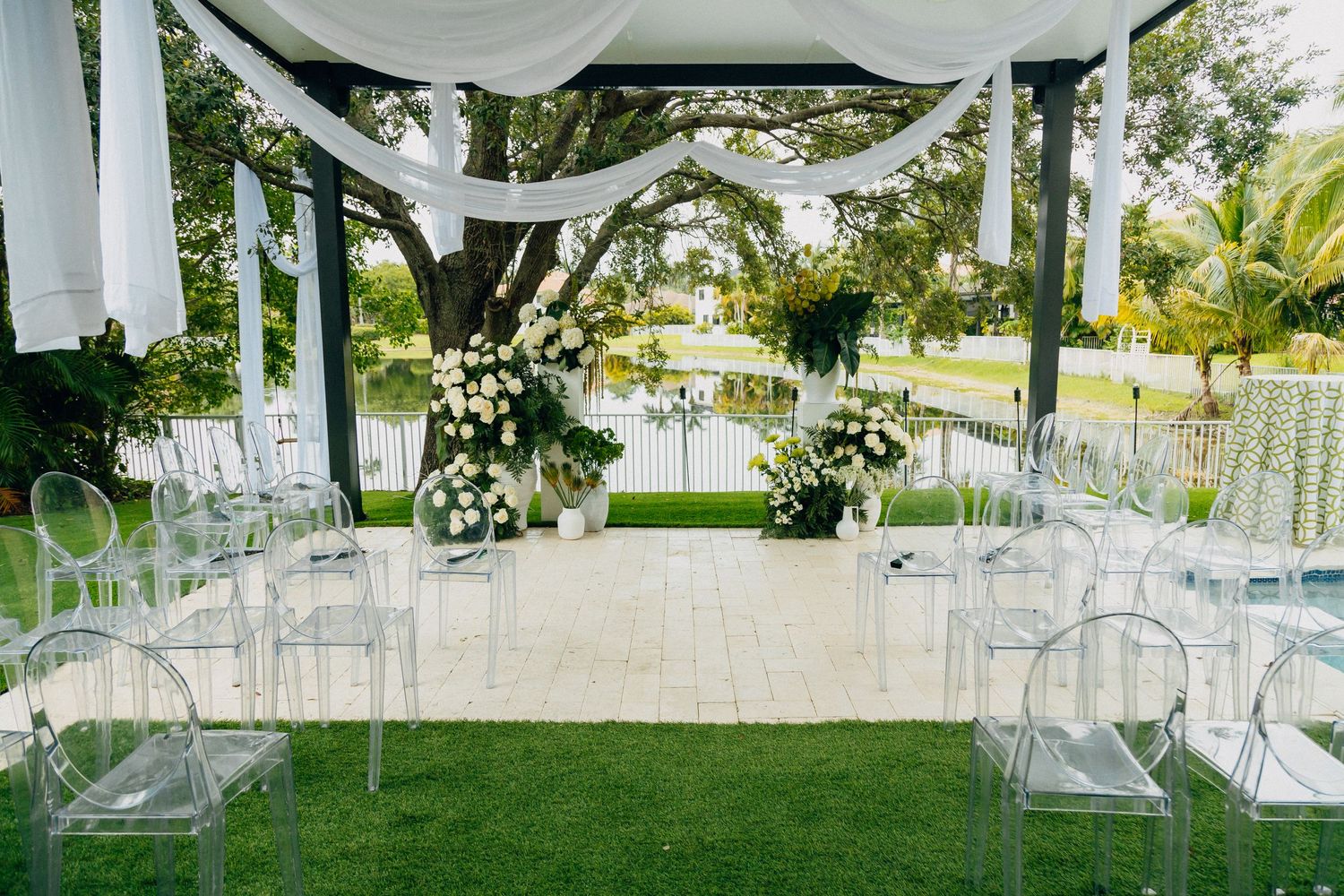Modern outdoor wedding ceremony setup with clear chairs and white fabric draping.