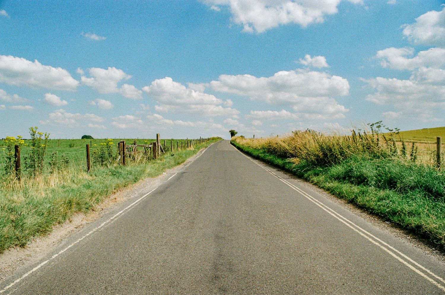 Empty rural road stretching into the distance between green fields under blue sky with white clouds.