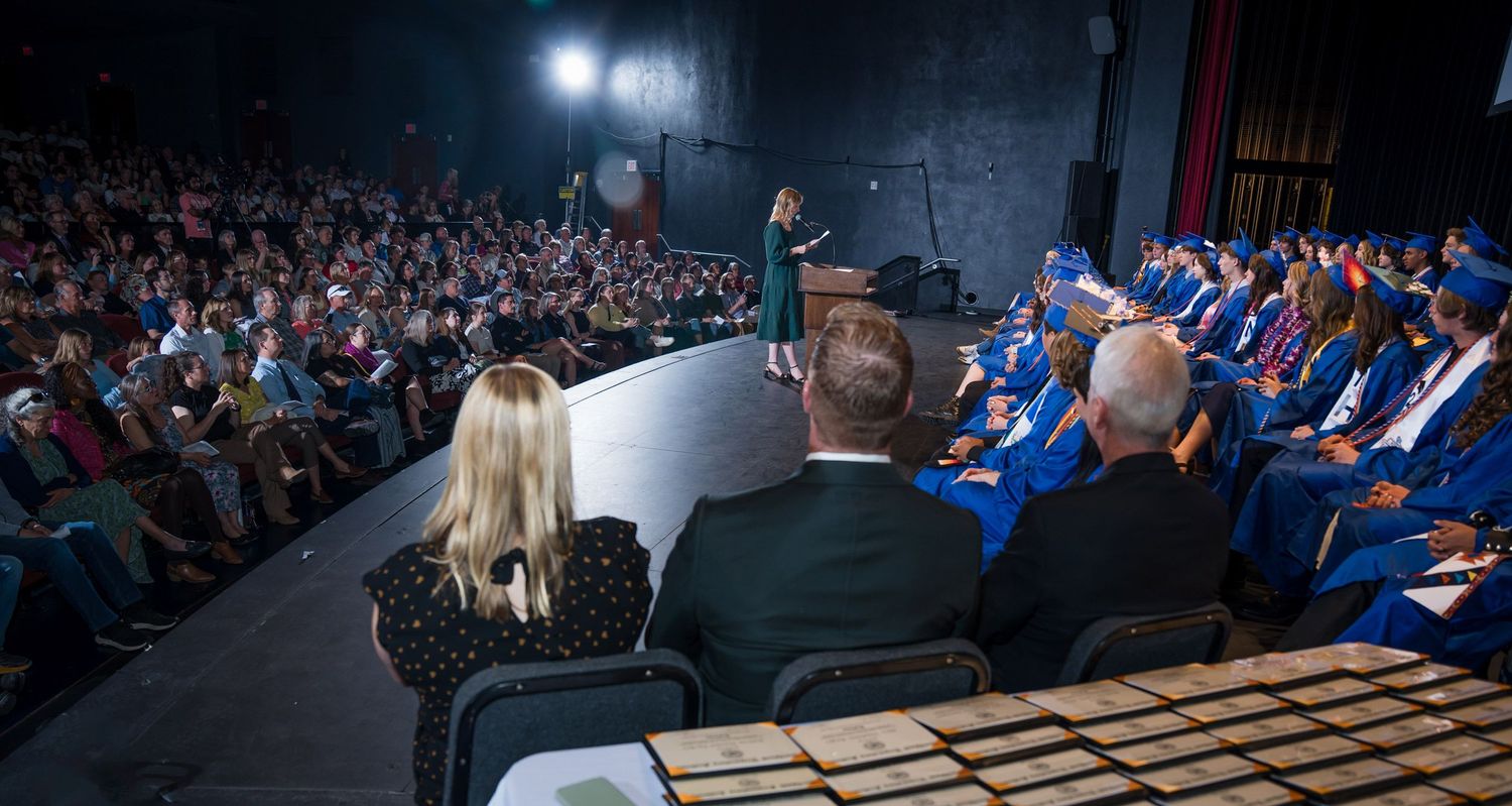 A large audience watches speakers on stage during a graduation ceremony in an auditorium with bright spotlights.