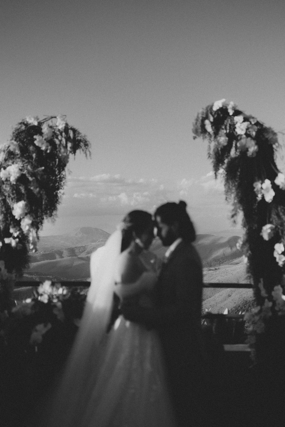 A romantic couple shares a kiss under floral arches against a scenic mountain backdrop at their outdoor wedding ceremony.