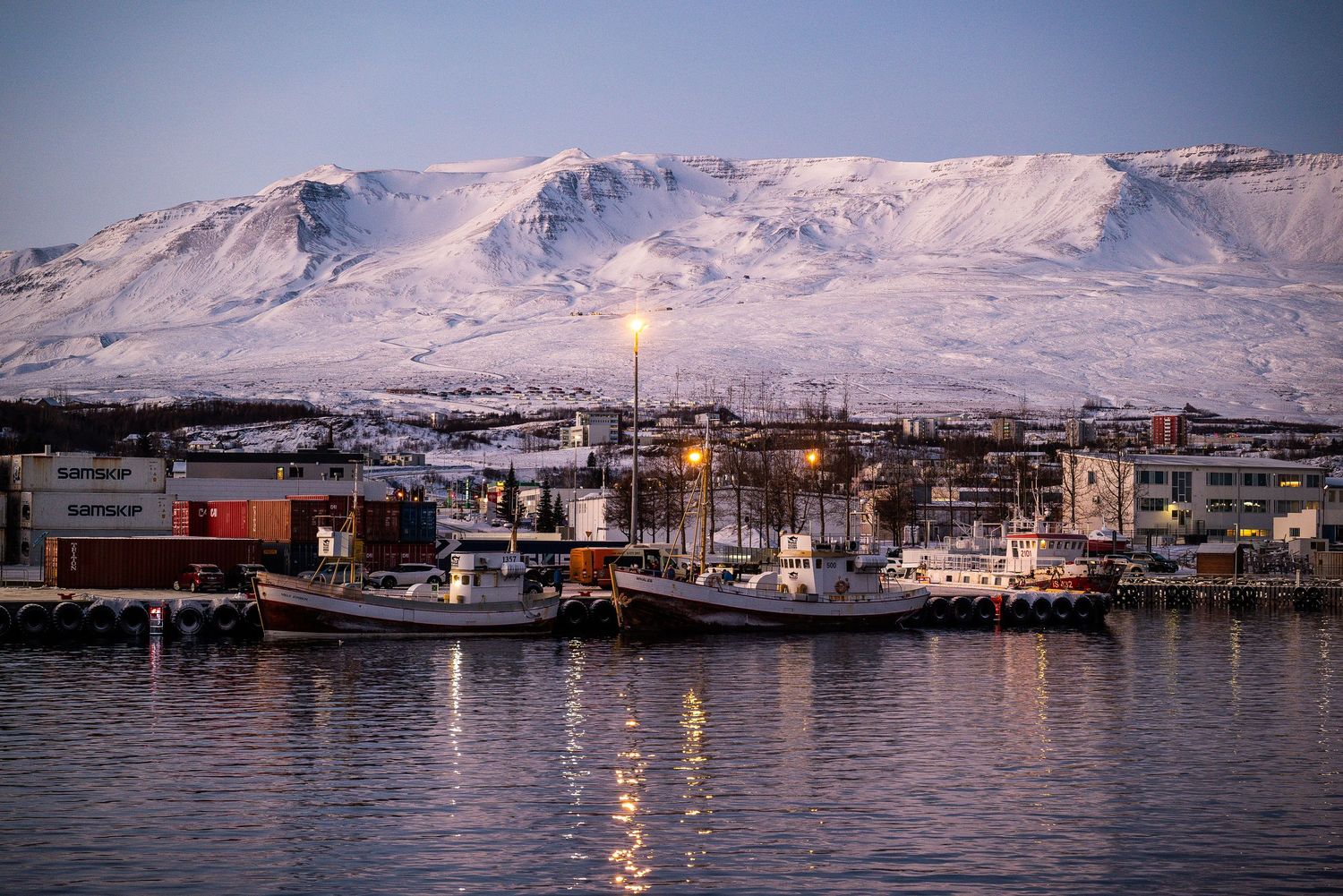 Harbor town with fishing boats lit at dusk beneath snow-covered mountain range.
