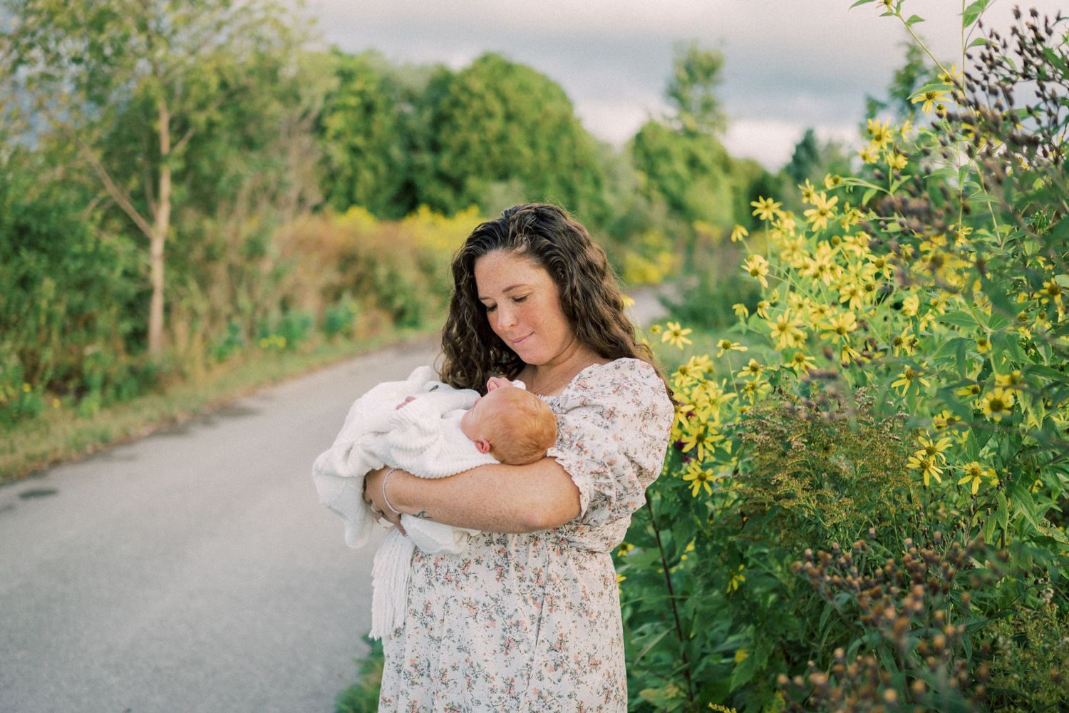 A tender moment on a country road with wildflowers as a woman in a floral dress cradles a newborn.