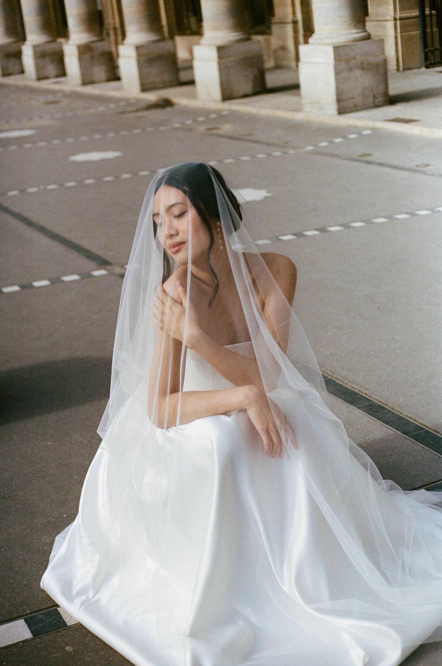A bride in a flowing white wedding dress and veil poses elegantly in an urban setting with pillared architecture.
