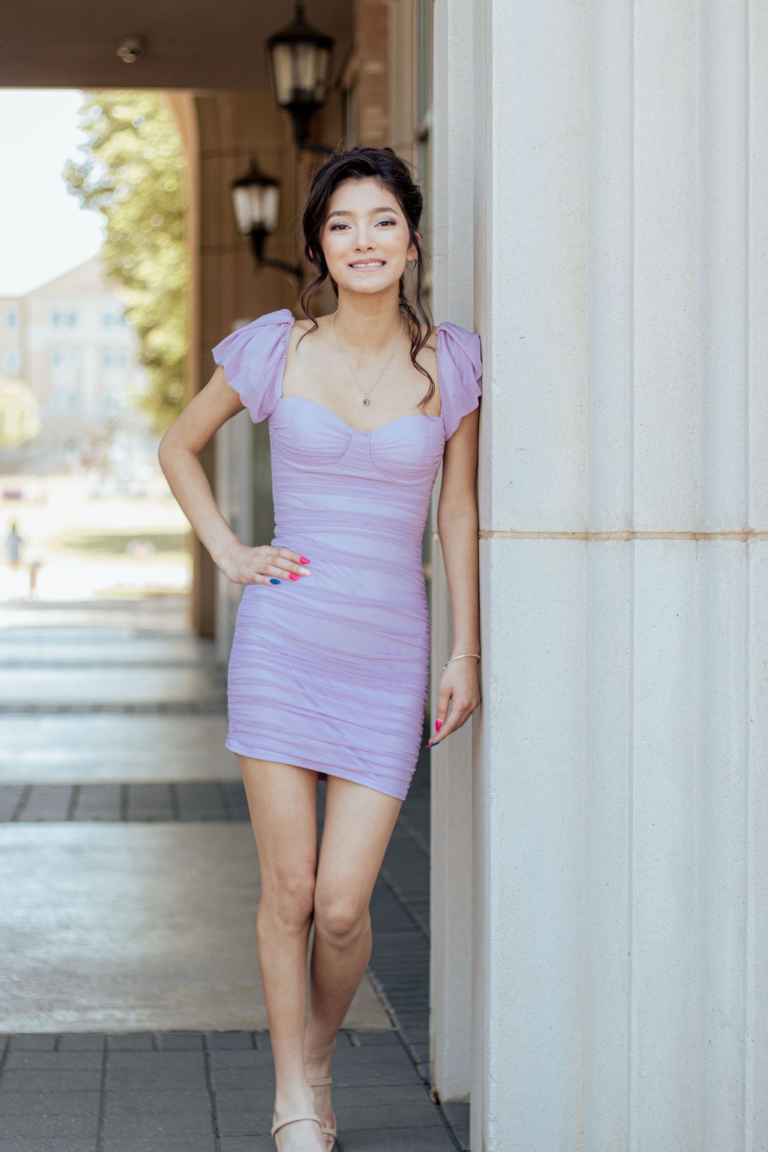 A person in a lavender-colored bodycon dress with cap sleeves poses in an outdoor covered walkway with lanterns.