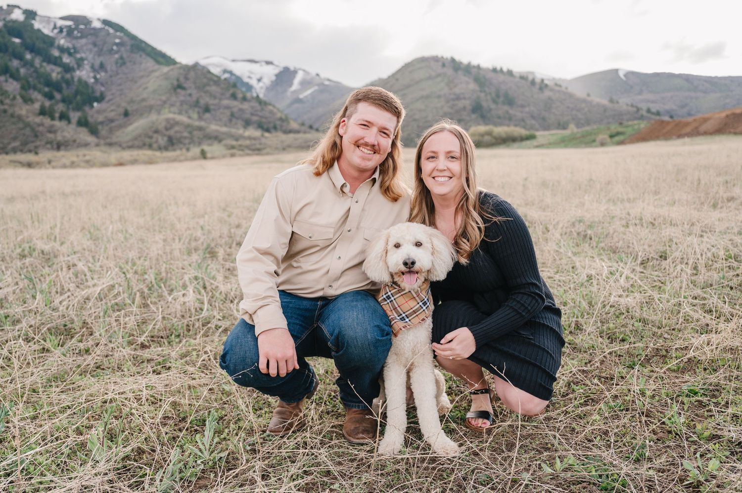 A couple posing with their white puppy in an open field with mountains in the background during autumn.