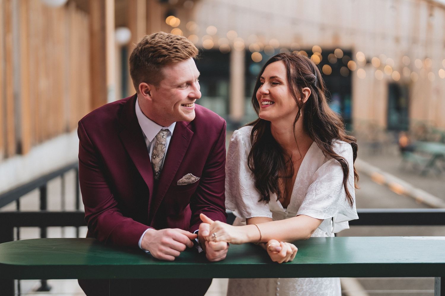 A couple in wedding attire pose happily against a dark gray wall, with the bride in white and groom in burgundy.
