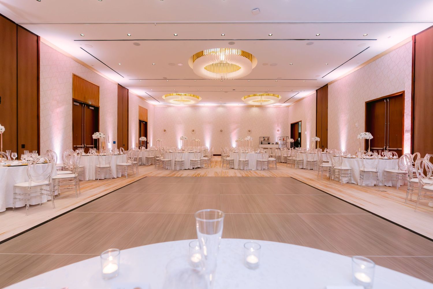 Elegant ballroom with white-themed decor, round dining tables, and crystal chandeliers for a wedding reception setup.