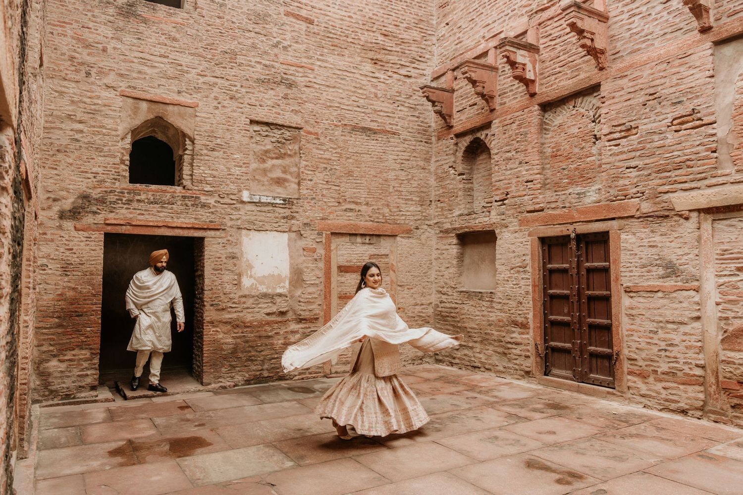 Two figures in traditional attire pose in a historical terracotta brick courtyard with rustic doorways and worn walls.