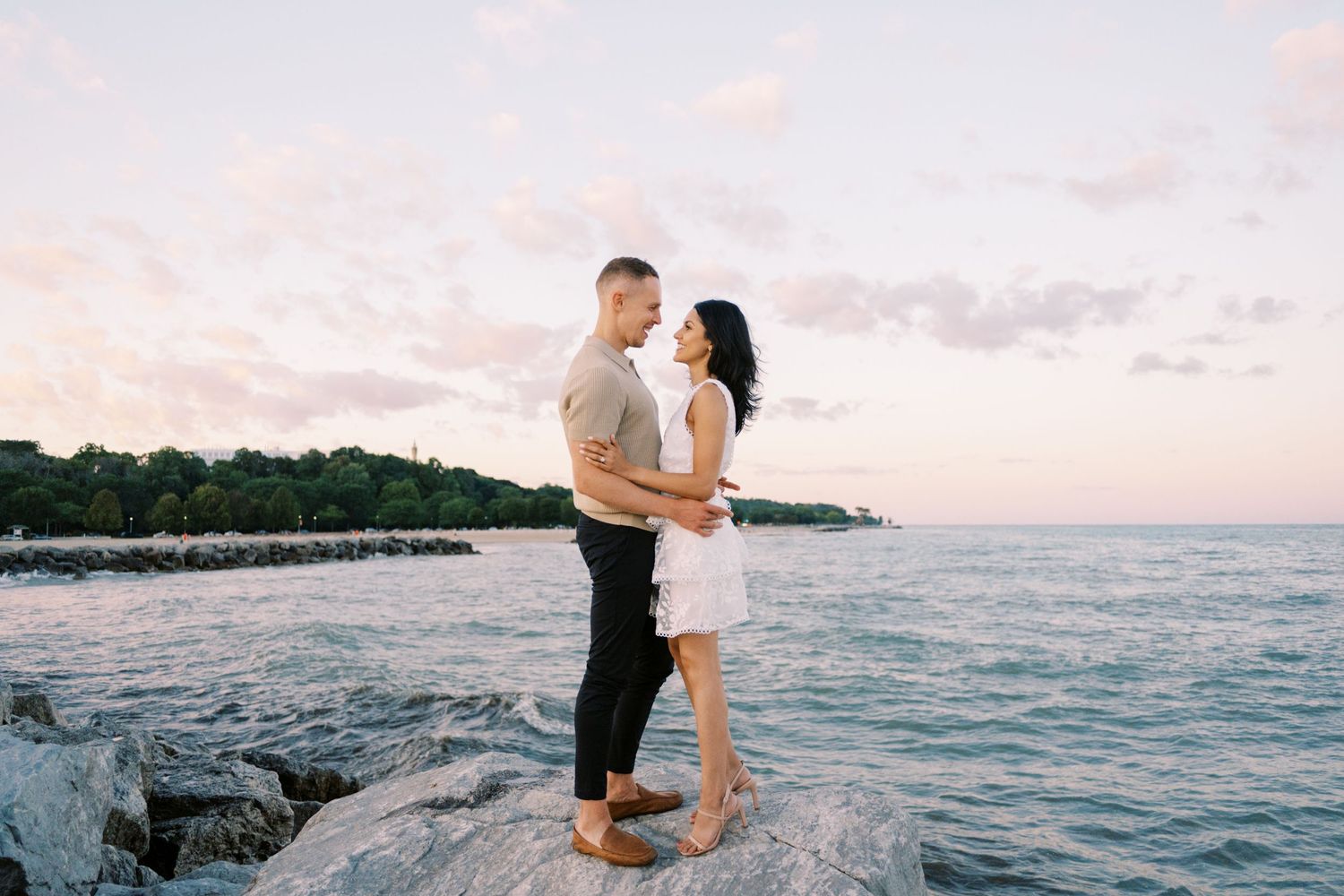 Couple embraces on rocky shoreline at sunset with ocean waves and pink sky in the background.