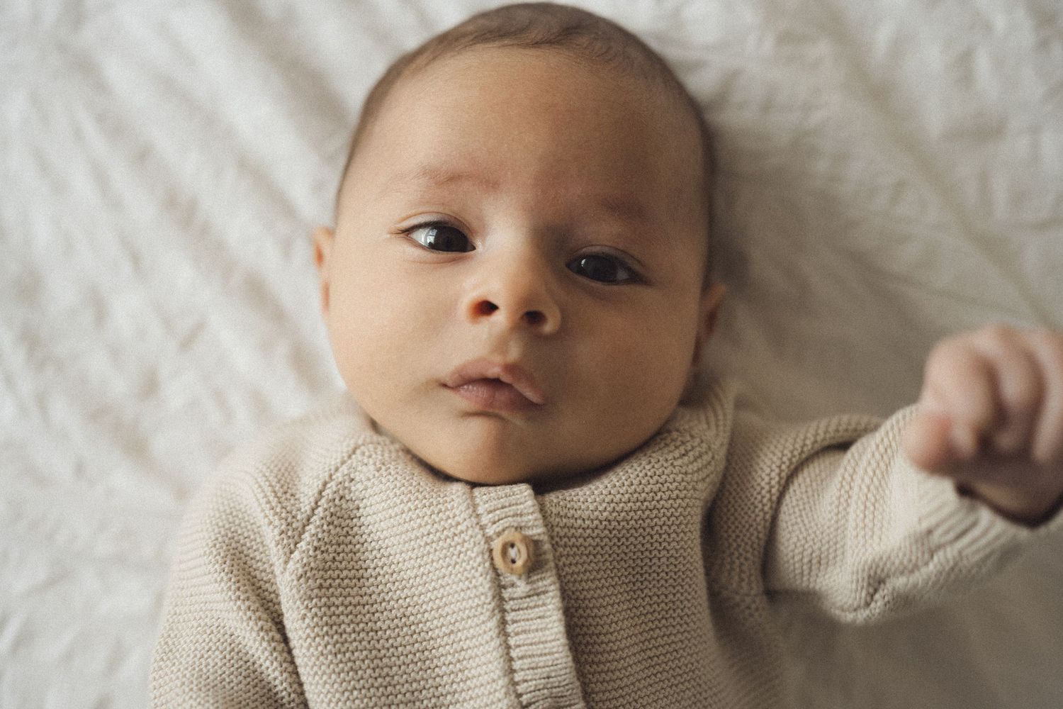 Baby in a cream knit sweater lying on white bedding with a thoughtful expression.